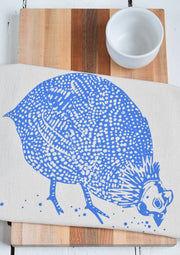 Hearth and Harrow - Organic Cotton Guinea Hen Tea Towel featuring a blue-violet hen design; showcased on a wooden surface beside ceramic kitchenware.
