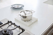 Yamazaki Home - Tower Folding Dish Drainer Mat, a white silicone trivet on a kitchen countertop, next to a stainless-steel pot, illustrating its versatile use.