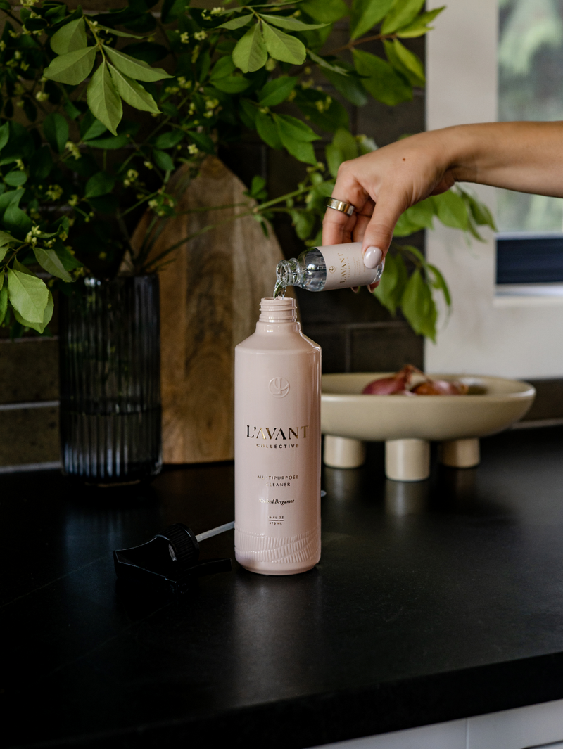 L'AVANT Collective Multipurpose Surface Cleaner in Blushed Bergamot scent, a hand pouring liquid into a luxury refillable bottle for versatile cleaning use.