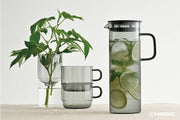 HARIO USA - HARIO Colors Glass Cold Brew Pitcher, 800ml: Grey, filled with ice water, lime, and mint, beside tinted-glass mugs and a vase with foliage.