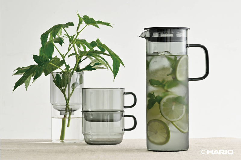HARIO USA - HARIO Colors Glass Cold Brew Pitcher, 800ml: Grey, filled with ice water, lime, and mint, beside tinted-glass mugs and a vase with foliage.