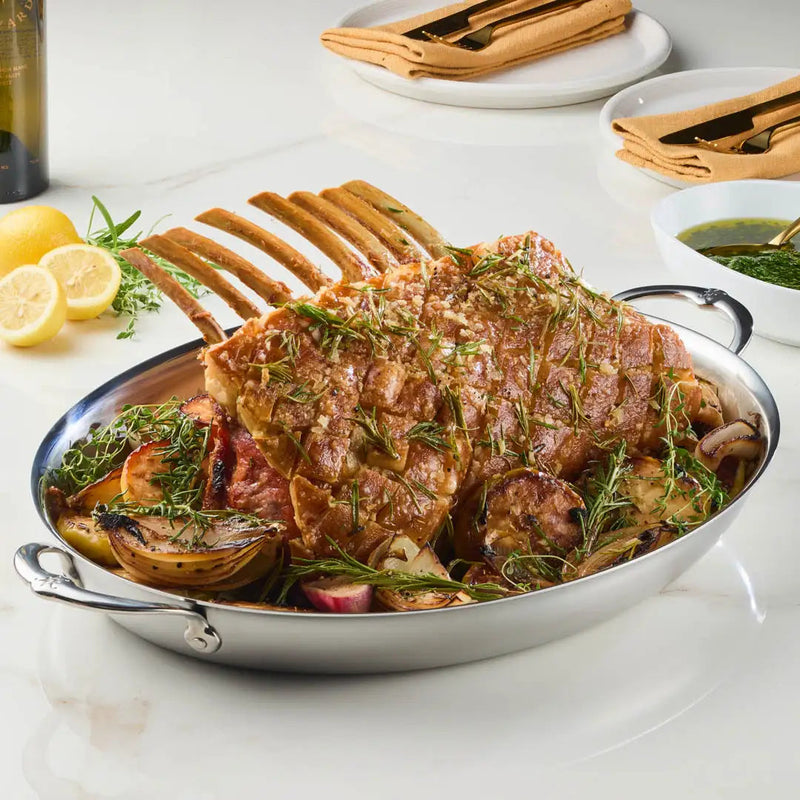 Hestan Provisions Large Clad Oval Roaster showcasing a roast rack of lamb on roasted vegetables, paired with herb sauce, lemons, and wine in a kitchen setting.