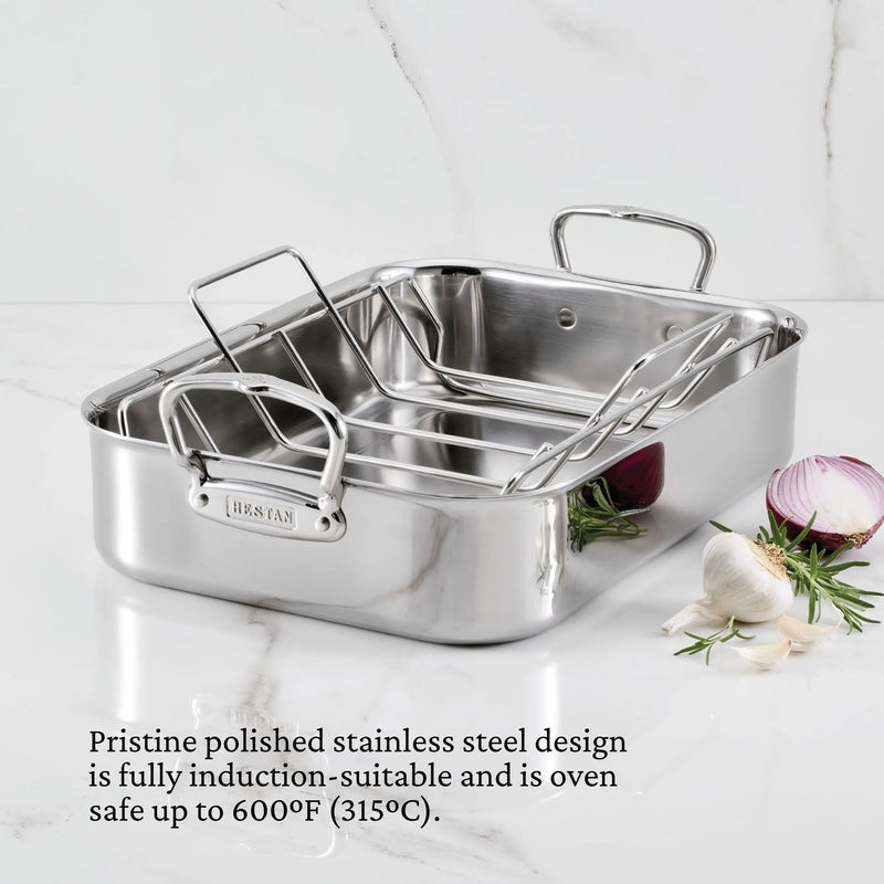Hestan | Polished Clad Stainless Steel Roaster with Rack, Small, on marble countertop, featuring heavy-duty handles and logo, surrounded by garlic, onion, and rosemary.