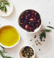 Kosterina - Greek Herb Kalamata Olives in a bowl, surrounded by oil, peppercorns, and herbs, perfect for salads or snacking.