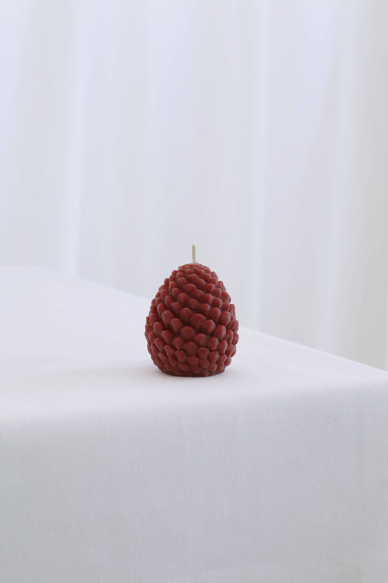 Mo&Co Home Exclusive: Sculpted Beeswax Pine Cones candle, red, pine-cone–shaped, hand-poured, on white cloth, evoking holiday nostalgia with contemporary elegance, available in two sizes.