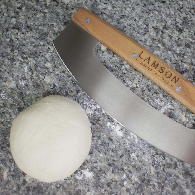 Lamson | 12 Pizza Rocker with Walnut Handles on a kitchen counter, next to a dough ball and knife, showcasing its precise cutting capability.