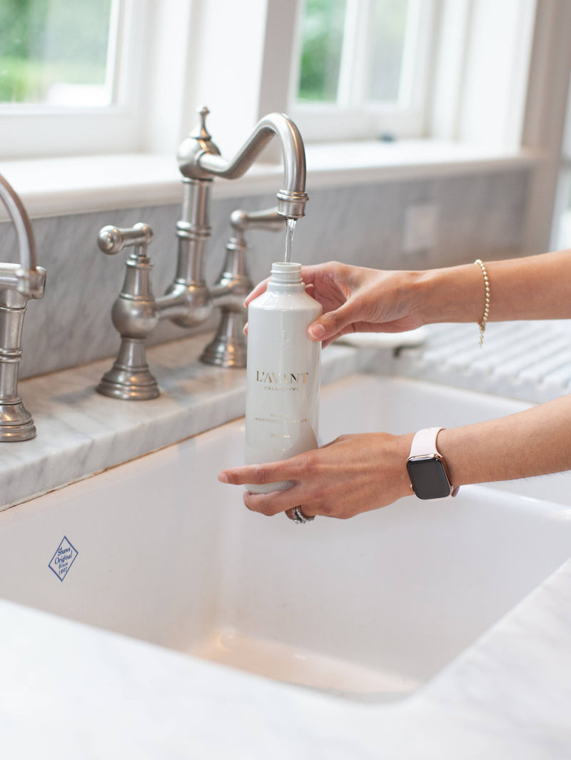 Person pouring water into L'AVANT Collective - Fresh Linen Multipurpose Cleaner Refill bottle, highlighting its eco-friendly refill process with glass packaging.