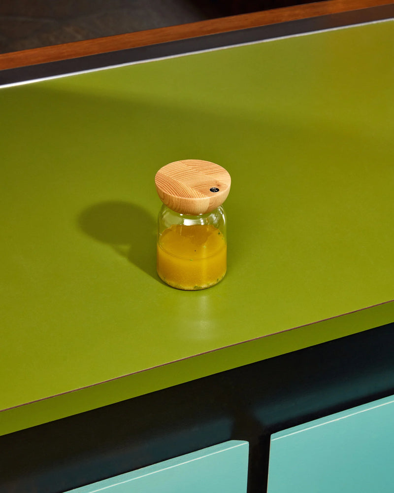 Holcomb Studio Totter Salad Dressing Shaker, a glass jar with a wooden lid, displayed on a green surface. Perfect for mixing and storing dressings.