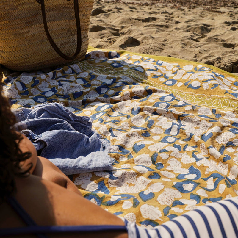 Couleur Nature - Forest Harvest Mustard & Blue tablecloth, 59 x 86, with floral patterns, lies on sand beside a woven tote, enhancing a beach setup.