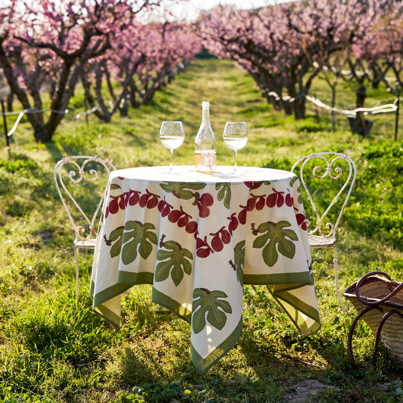 Couleur Nature - Fig Red & Green French Tablecloth drapes a bistro table in an orchard, featuring a fig leaf pattern, wine glasses, and garden chairs.