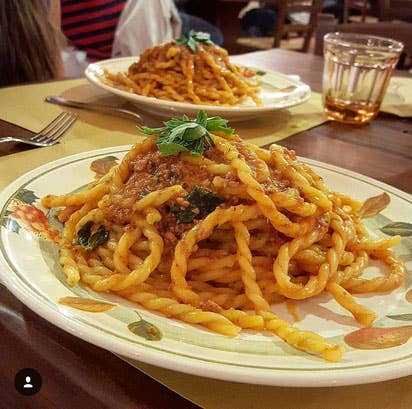 Two plates of Zia Pia - Fusilli Della Regina by La Fabbrica della Pasta, dressed in tomato sauce, garnished with herbs, on a wooden table in a casual restaurant setting.