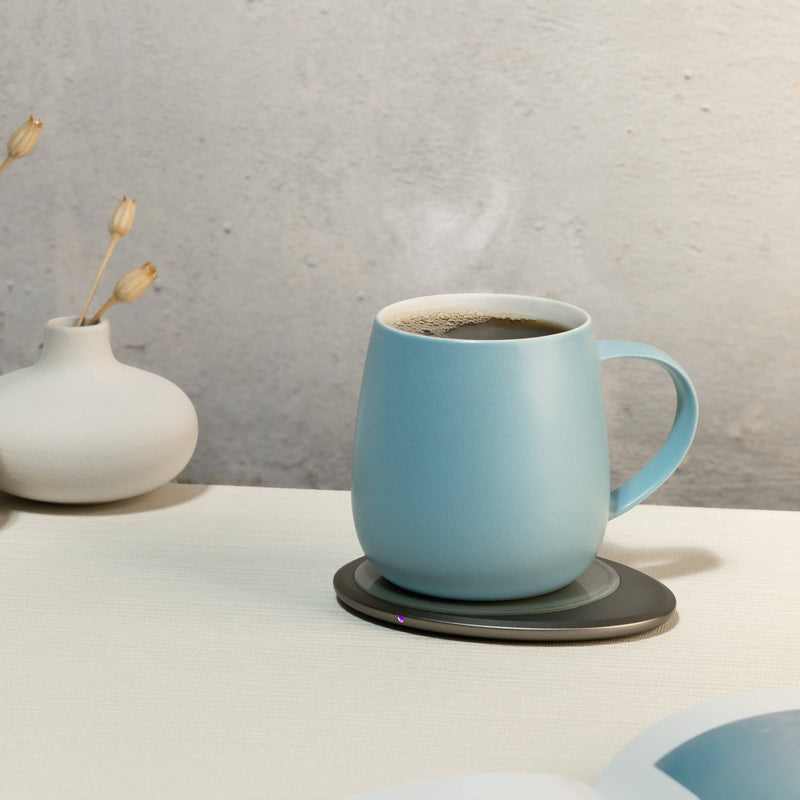 OHOM - Ui 3 Self-heating Mug Set: Moonmist Blue on an electric coaster, with a bud vase and open book nearby, creating a serene tabletop scene.