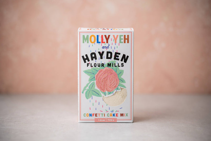 Hayden Flour Mills - Molly Yeh x HFM Confetti Cake Mix box featuring a rose graphic and sprinkles, photographed against a pink–peach background.