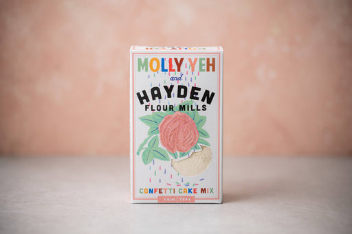 Hayden Flour Mills - Molly Yeh x HFM Confetti Cake Mix box featuring a rose graphic and sprinkles, photographed against a pink–peach background.