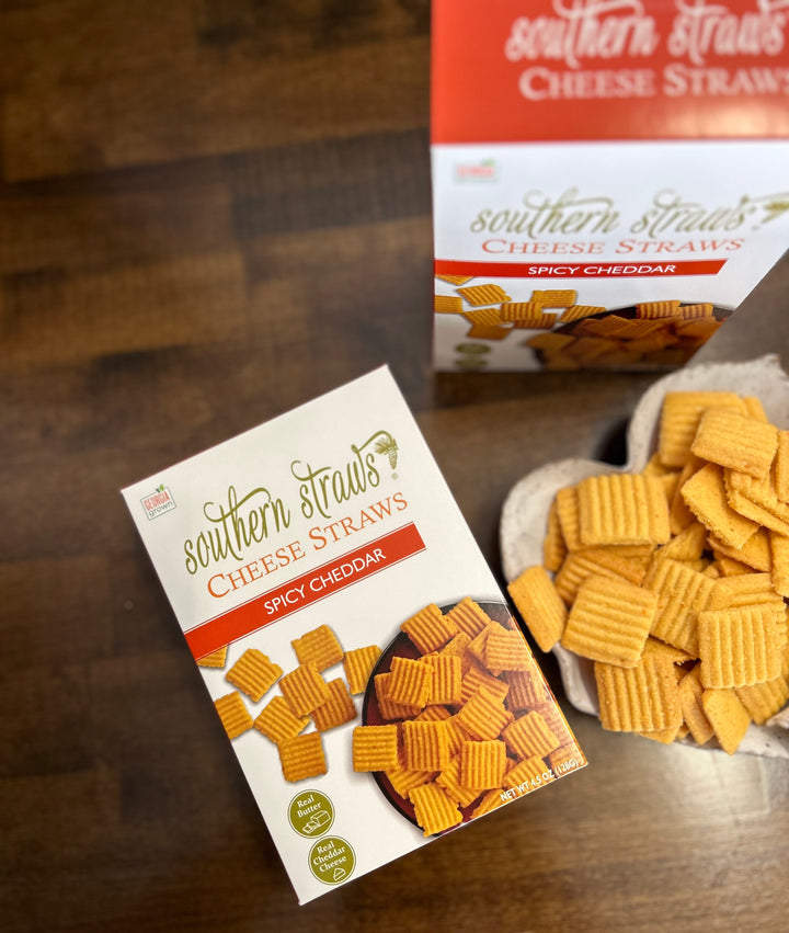 Southern Straws | Cheese Straws: Box of Spicy Cheddar cheese straws beside a bowl of ridged crackers, showcasing handcrafted, spicy Southern snack.