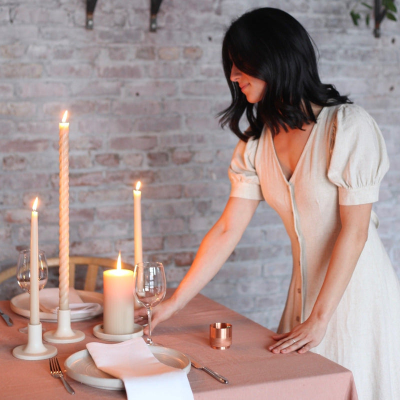 Woman arranges a candlelit dinner table featuring The Floral Society | Match Strike and Holder, a copper vessel for strike anywhere matches, on a muted rose-beige tablecloth.