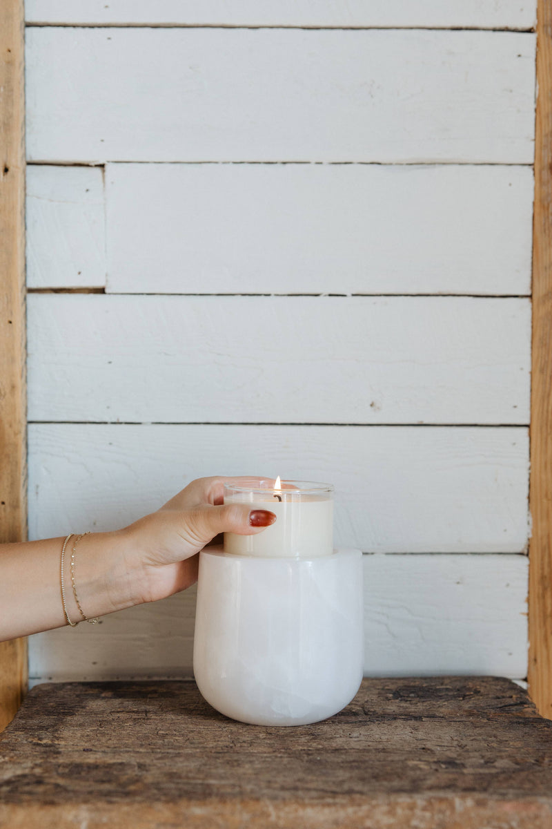 Hand with red nails places a lit Last Candle Co - White Onyx Refillable Candle into a marble vessel on a rustic wooden table.