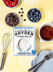 A breakfast-themed flat-lay featuring Hayden Flour Mills White Sonora Pancake Mix, surrounded by raspberries, blueberries, chocolate chips, maple syrup, a whisk, and plates with forks.
