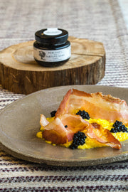 TartufLanghe Truffle Perlage pearls enhance creamy scrambled eggs with black caviar and crispy cured pork, showcased on a rustic plate alongside the product jar.