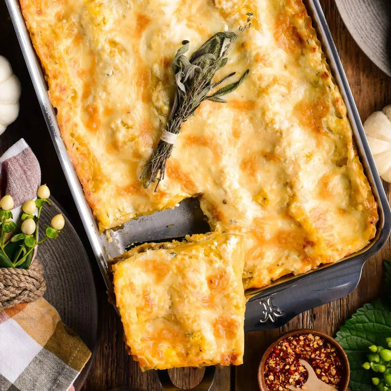 Hestan | Oven Bond Tri-Ply Bakeware, Rectangular Baker holding a cheesy, layered casserole with a portion removed, showing golden crust and herb garnish on rustic table.