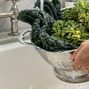 Hestan Provisions | Stainless Steel Colanders, 5 Quart, efficiently rinsing kale and broccolini under a faucet, showcasing its superior drainage and durability in kitchen tasks.