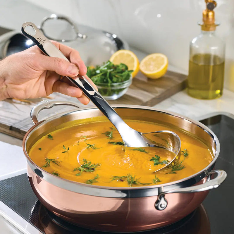 Hestan Provisions | Stainless Steel Ladle, 13 (5oz) lifting herb-garnished orange soup in a modern kitchen.