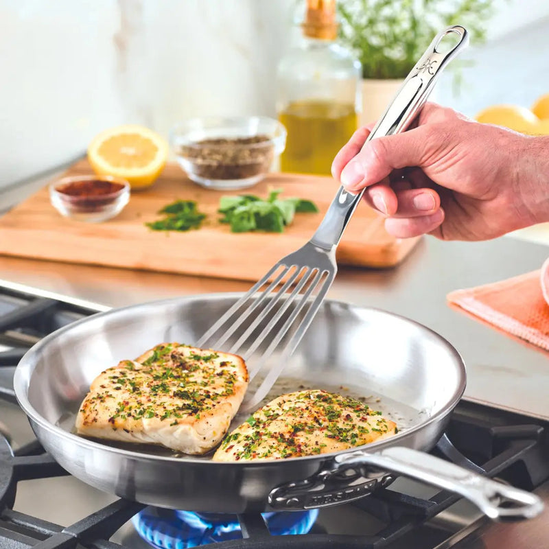 Hestan | Stainless Steel Flexible Fish Spatula, 13 flipping herb-seasoned chicken in a stainless-steel pan over a blue flame, amidst spices and a cutting board.