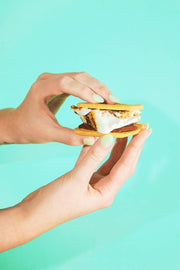 Hands with mint-green nails holding a s'more made with Malvi Mallow - Honey Vanilla Marshmallows, showcasing the gooey marshmallow between cookies and chocolate.