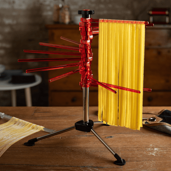 Marcato Tacapasta Pasta Drying Rack with red arms, holding freshly made pasta sheets on a wooden countertop. Includes removable rod for cutting and collecting pasta.