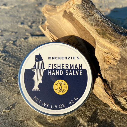 MacKenzie's Fisherman Hand Salve (1.5 oz) tin on sandy ground with driftwood; all-natural, highly moisturizing, perfect for dry skin, made in New England.