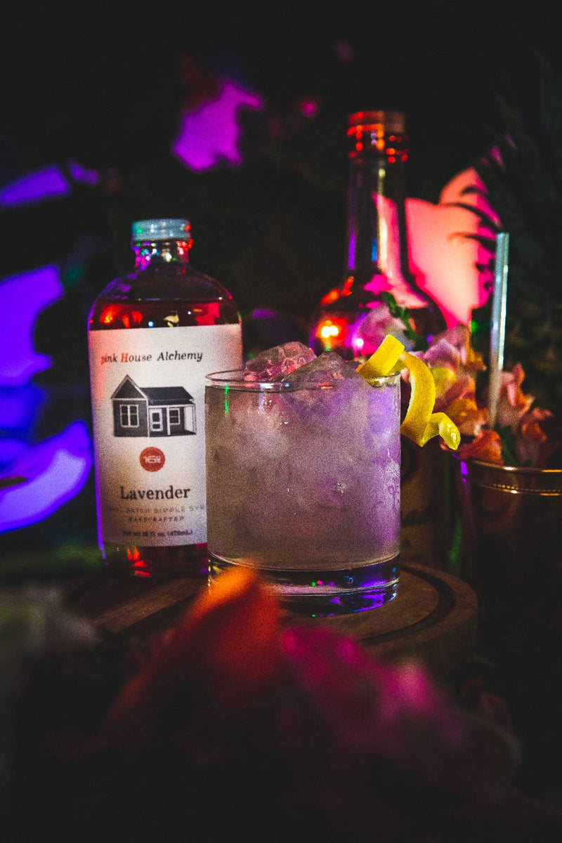 Pink House Alchemy - Lavender Simple Syrup bottle with a house illustration, next to a glass of iced lavender drink with lemon.