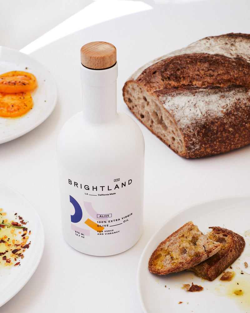 Brightland - ALIVE Extra Virgin Olive Oil with artisan sourdough, ripe tomato slices, and seasoned olive oil on a bright white table setting.