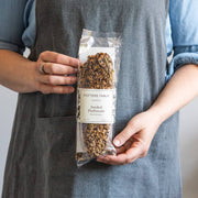 Person in a grey apron holds Potters Table Seeded Olive Oil Flatbreads, packaged to showcase grain- and seed-topped crackers, emphasizing handcrafted quality and award-winning taste.