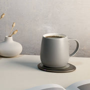 OHOM - Ui 3 Self-heating Mug Set: Jasmine White with steaming coffee on a warming coaster, alongside a white vase with dried buds and an open book corner.