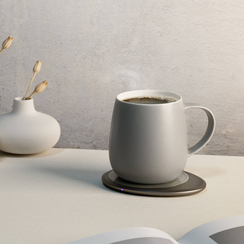 OHOM - Ui 3 Self-heating Mug Set in soft gray, on a warming plate, with a sculptural vase and open book, enhancing your coffee experience.