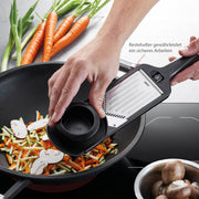 Hand using Microplane Adjustable V-Blade Mandoline Slicer to julienne carrots and zucchini into a wok on an induction cooktop, surrounded by fresh vegetables.