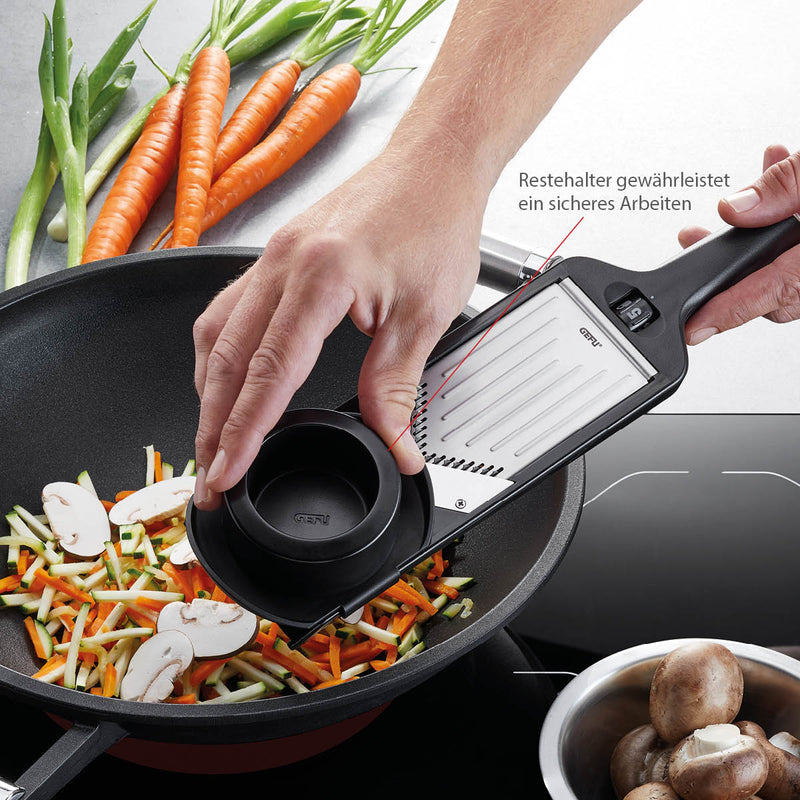 Hand using Microplane Adjustable V-Blade Mandoline Slicer to julienne carrots and zucchini into a wok on an induction cooktop, surrounded by fresh vegetables.