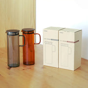 HARIO USA - HARIO Colors Glass Cold Brew Pitcher, 800ml: Grey and amber pitchers with mesh filters, elegantly presented in matching retail boxes on a wooden tabletop.