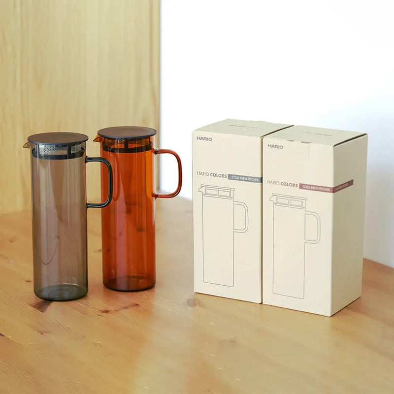HARIO USA - HARIO Colors Glass Cold Brew Pitcher, 800ml: Grey and amber pitchers with mesh filters, elegantly presented in matching retail boxes on a wooden tabletop.