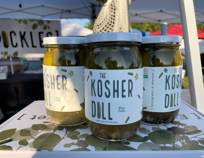Three jars of PICKLES by Evergreen and Market - The Kosher Dill - Spears, displayed at an outdoor market, showcasing whole pickles with spices and garlic in brine.