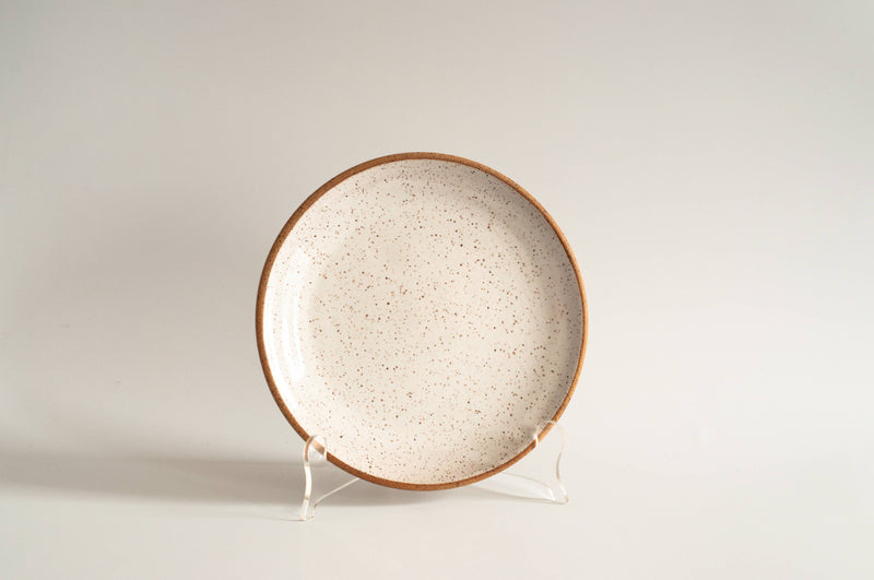 RachaelPots - Salad Plate: Coral. White ceramic plate with brown speckles and rim, displayed on a clear stand. Hand-thrown, microwave and dishwasher safe, 8.5 diameter.