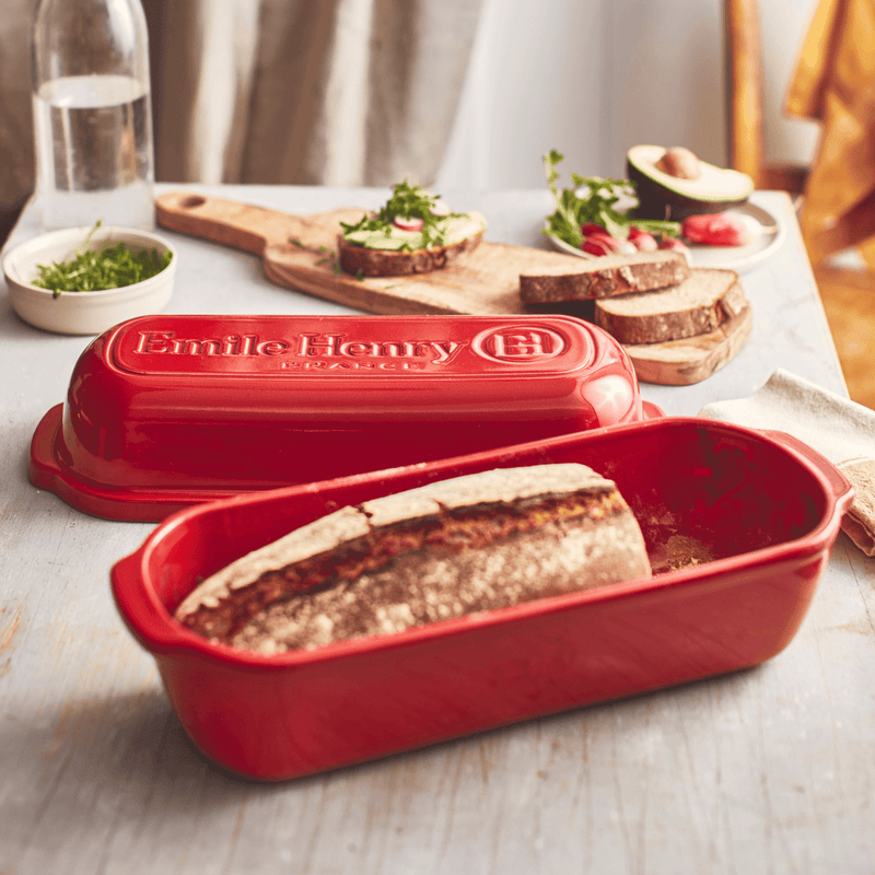 Emile Henry Loaf Bread Baker with freshly baked crusty bread on a wooden tabletop, surrounded by sliced bread, avocado toasts, and kitchen essentials.