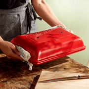 Person lifting lid of Emile Henry | Baguette Baker, Burgundy, on wooden surface with cutting board and knife, showcasing its design for baking crusty baguettes.