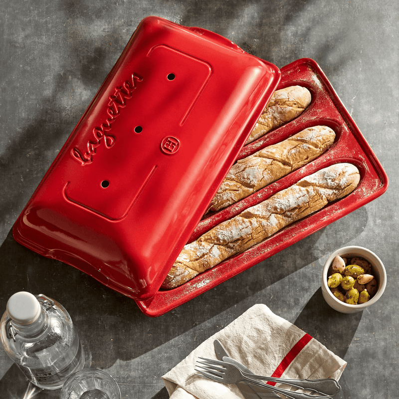 Emile Henry | Baguette Baker, Burgundy holds three baked baguettes. Features a lid for perfect humidity. Includes olives, garlic, water, and cutlery setting.
