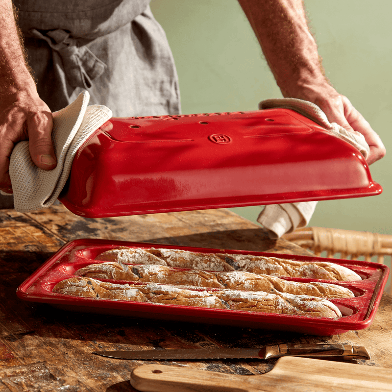 A person holds an Emile Henry Baguette Baker, showcasing a fresh loaf inside the red pan designed for perfect home-baked baguettes.