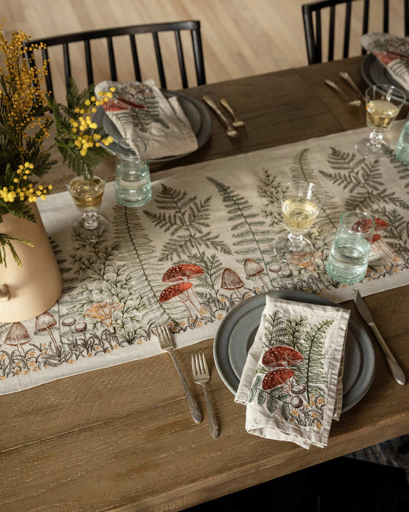 Coral & Tusk - Mushrooms and Ferns Bouquet Dinner Napkin displayed on a rustic table setting, complementing the table runner with matching botanical motifs.