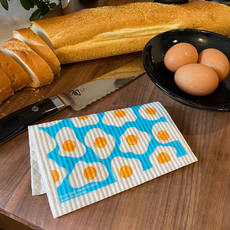 Bespoke Provisions - Eggs Swedish Dishcloths displayed beside bread and eggs on a table, highlighting their versatility for cleaning kitchen surfaces effectively.