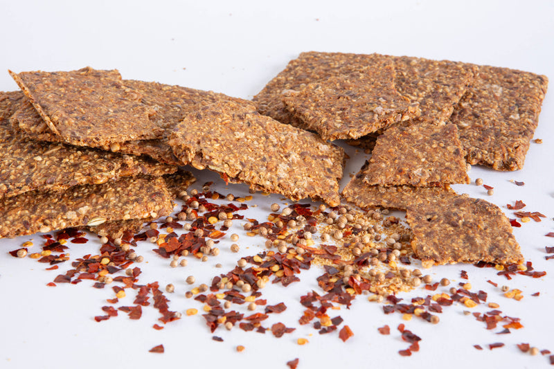 Norwegian Baked - Chili & Coriander Knekkebrød crispbread, featuring whole-grain and seed textures, with scattered chili flakes and coriander seeds on a white surface.