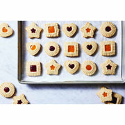 King Arthur Thumbprint Cookie Cutters 4-Piece Set on a baking sheet, showcasing embossed cookies with jam-filled centers in various shapes: hearts, stars, squares, and rounds.