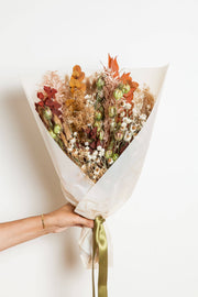 Idlewild Floral Co. - Fall Dried Flower Bouquet: A hand holds a warm-toned bouquet of dried flowers, wrapped in paper with a satin ribbon, ideal for seasonal home decor.